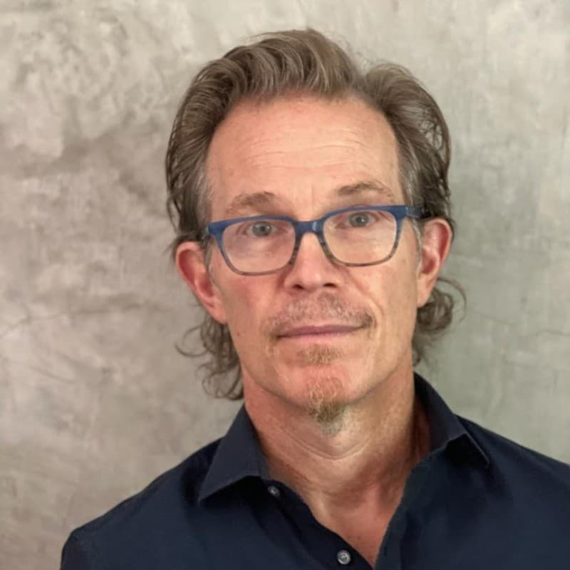 Man with wavy hair, blue glasses, and a goatee against a textured gray wall.