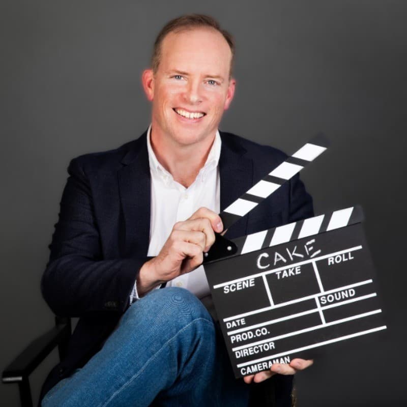 Smiling man in a blazer holding a movie clapperboard with "CAKE" written on it.
