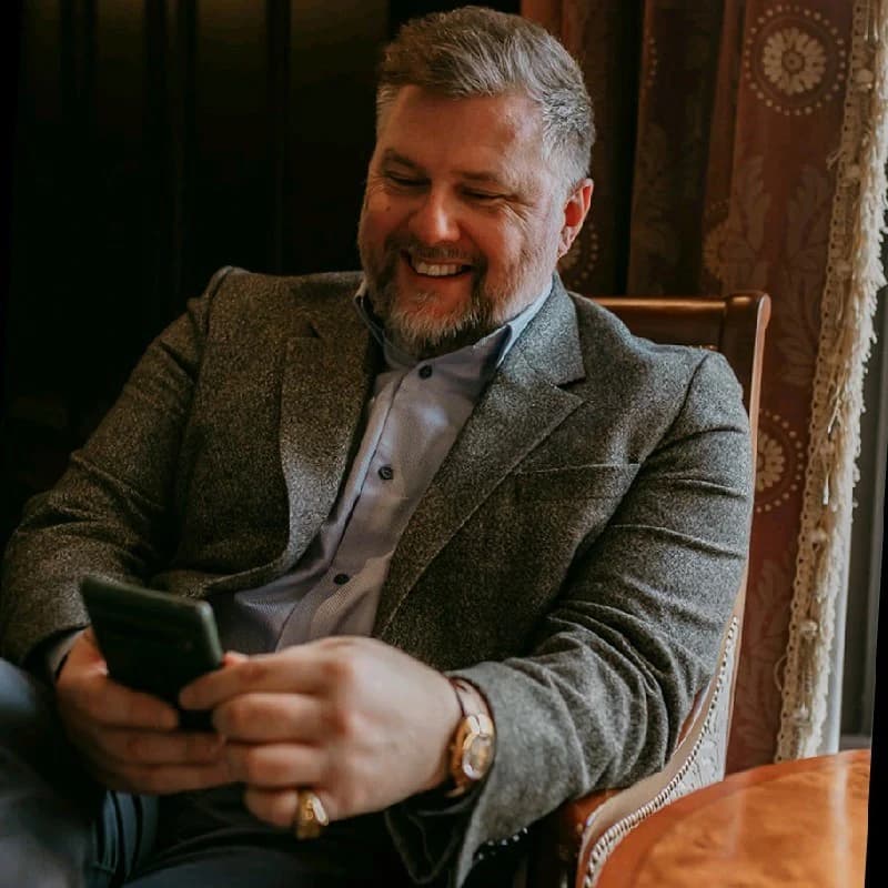 Bearded man in a grey blazer smiling warmly while looking at his smartphone screen.